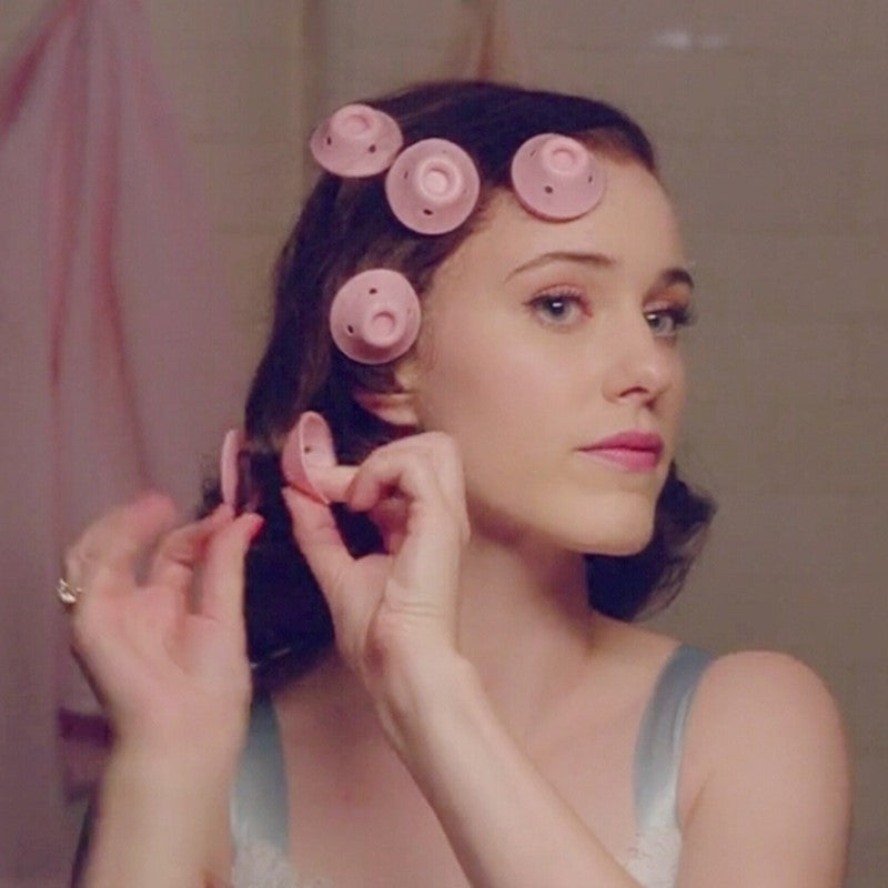 Woman with hair in pink rollers applying makeup in a bathroom.